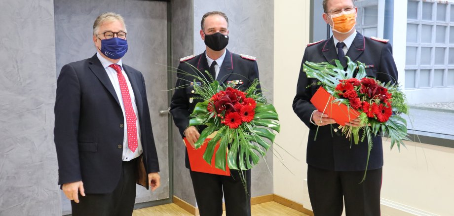 Drei Männer stehen nebeneinander. Die Herren mittig und links sind von der Feuerwehr und halten Blumensträuße in der Hand. Alle drei Tragen einen Mund-Nasen-Schutz.