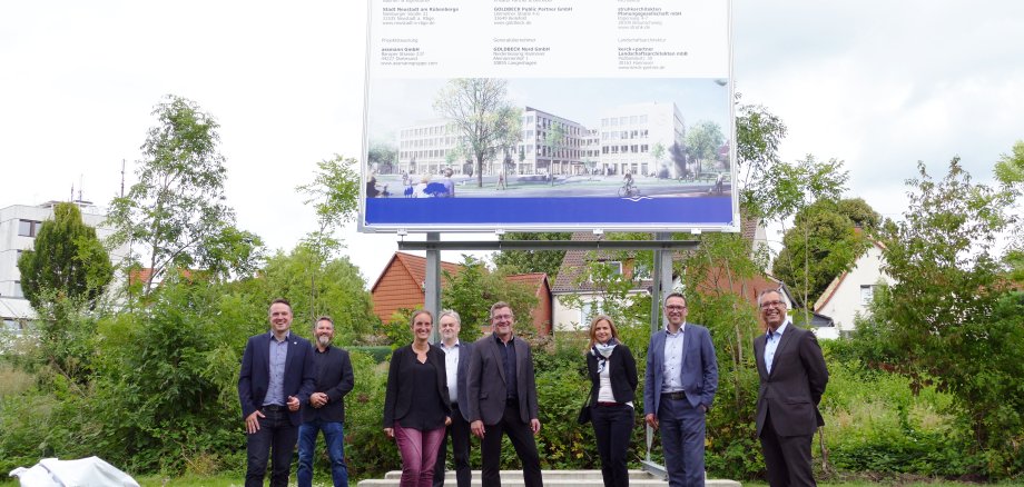Acht Personen stehen auf einer Grünfläche vor einem rund vier meter hohem Baustellenschild. Dies zeigt den Rathausneubau und die beteiligten Firmen. 